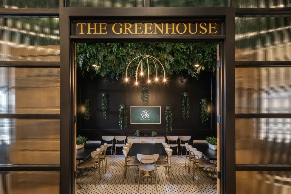 View through the entrance of The Green House at Wire Park into a room with tables, chairs, greenery, and hanging lights.