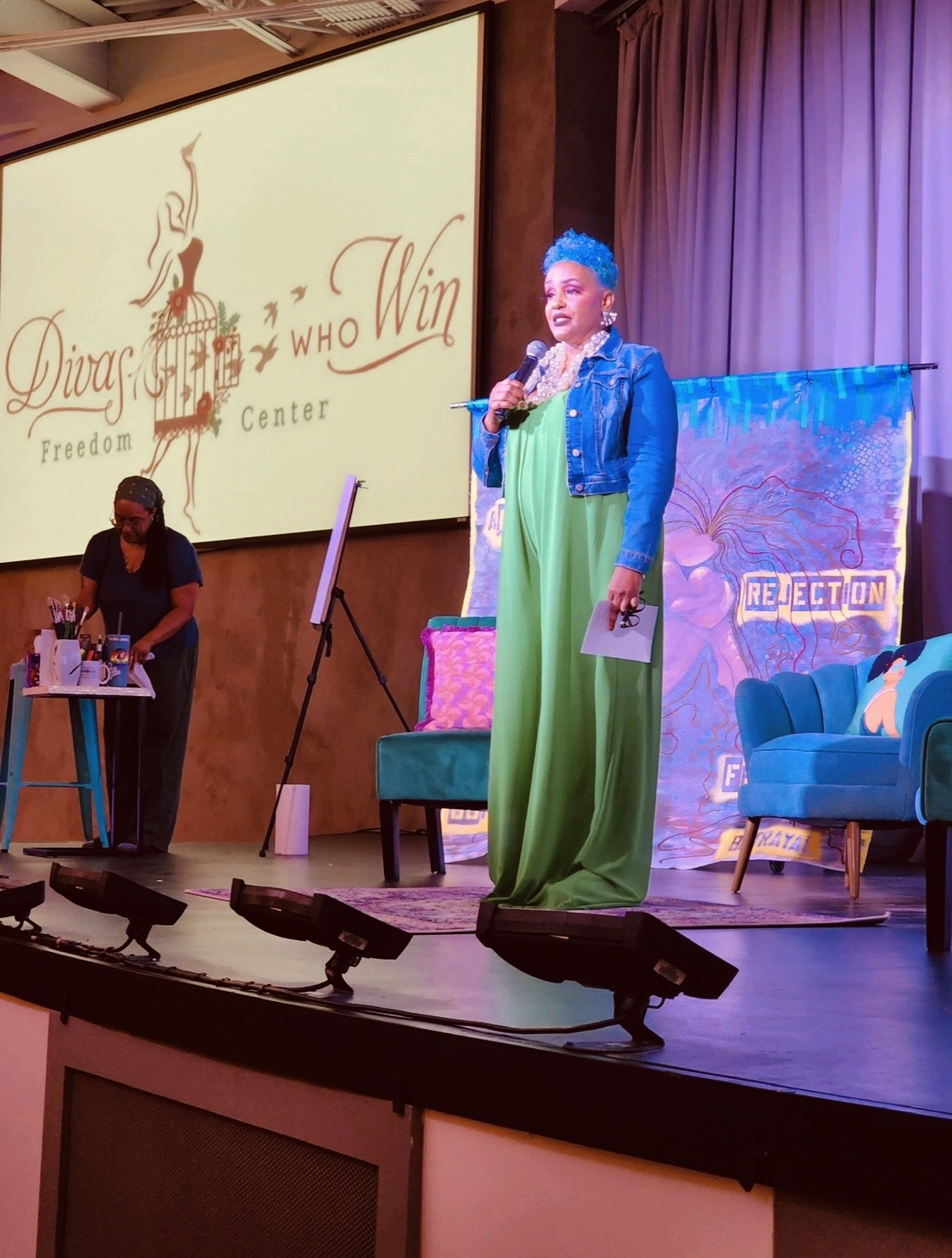 Chanda Santana speaking on stage beside a Divas Who Win art installation