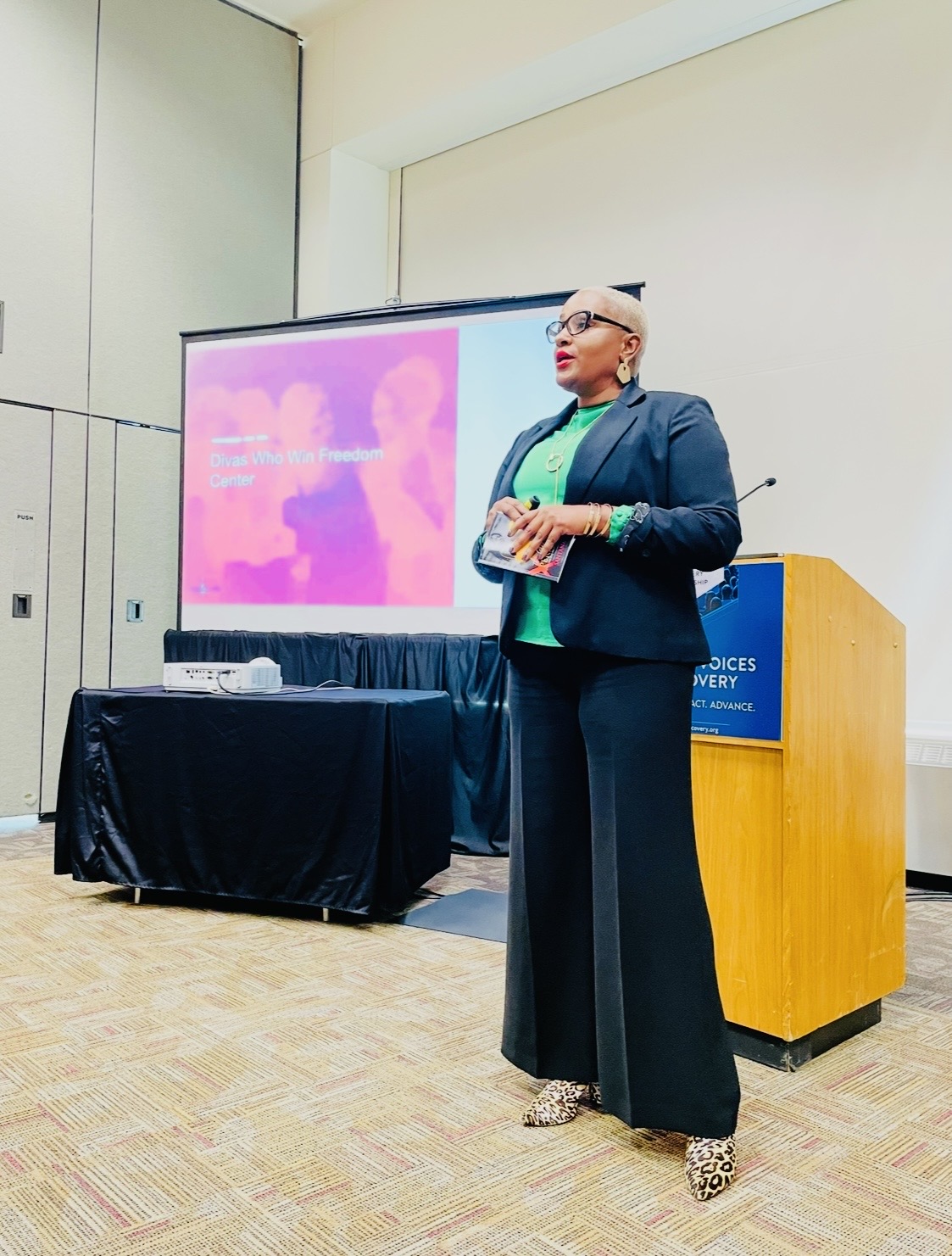 Chanda Santana presenting beside a conference podium and a Divas Who Win Freedom Center slide